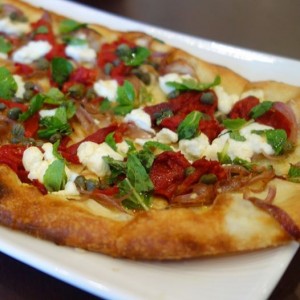 The dough is made in-house, layered with roasted tomatoes, caramelized onions, capers, mint and goat cheese, drizzled with olive oil, then baked in our pita oven. 